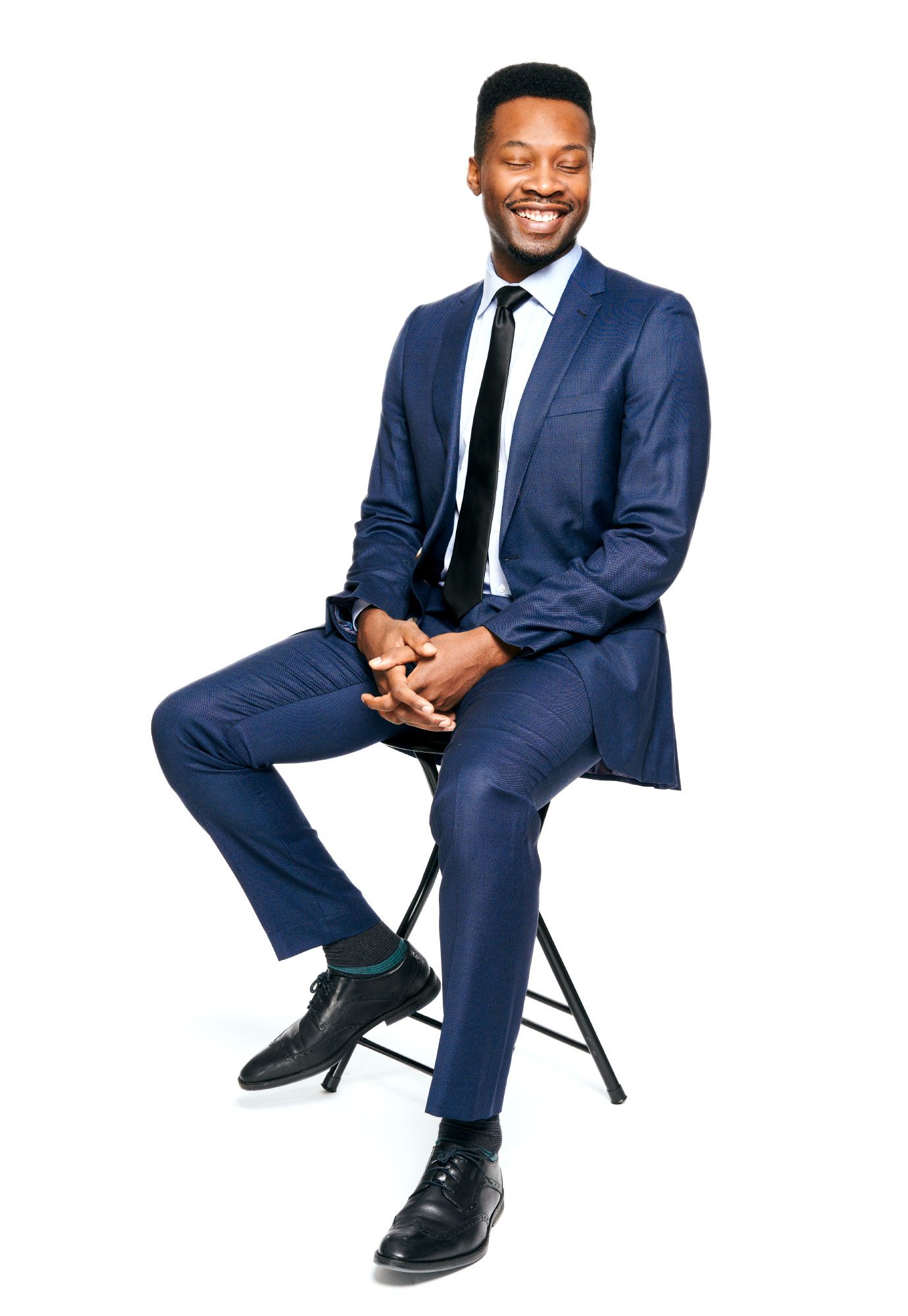 Smiling businessman in navy blue suit sitting on stool against white background.