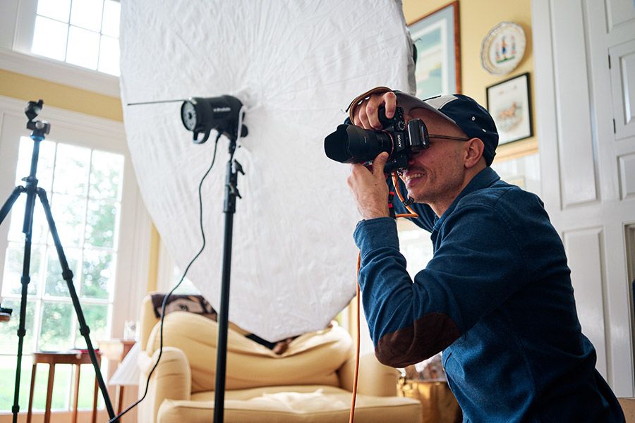 A professional photographer uses a DSLR camera with studio lighting equipment set up in a warmly decorated living room.