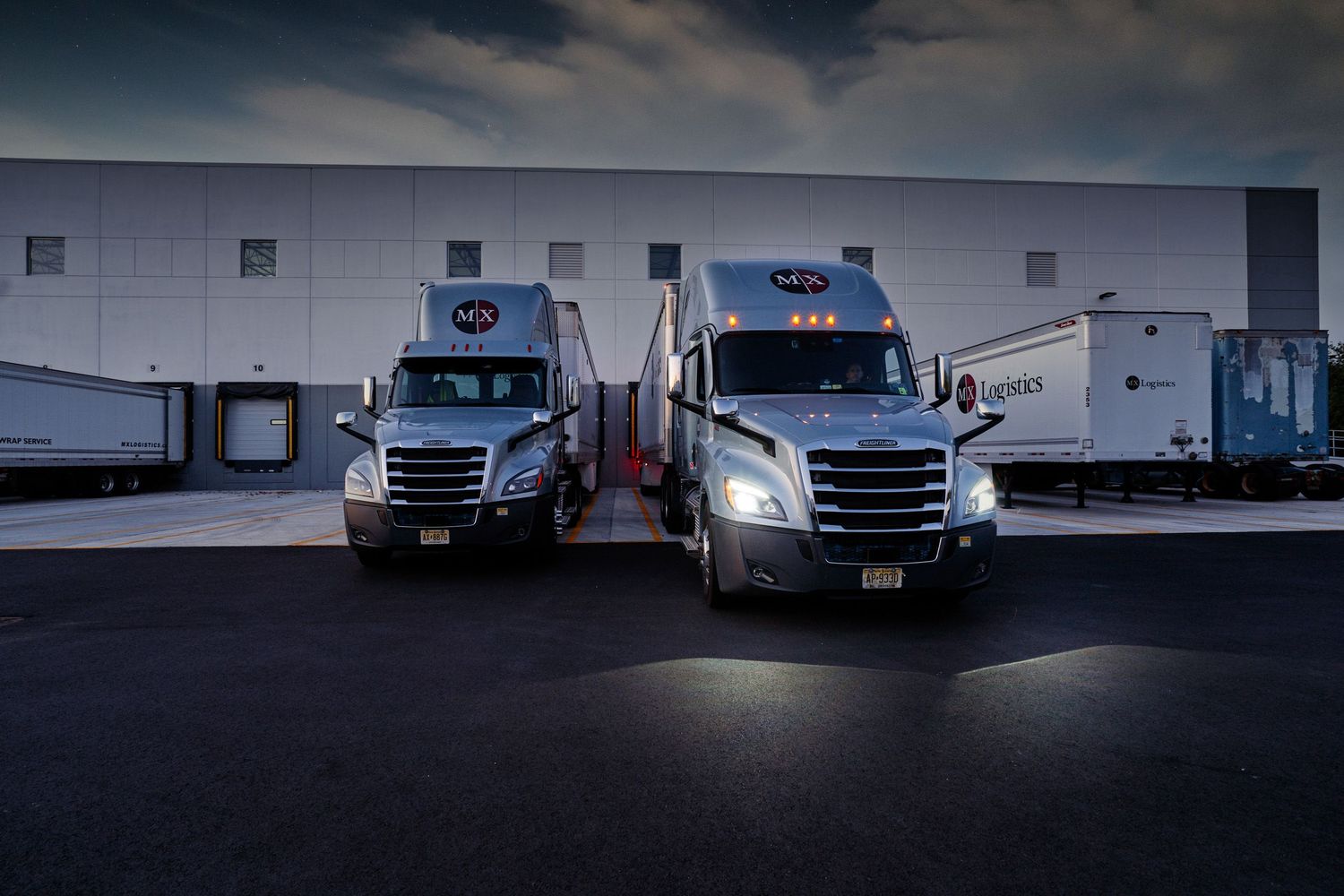 Two red semi-trucks parked at a logistics warehouse loading dock at dusk.