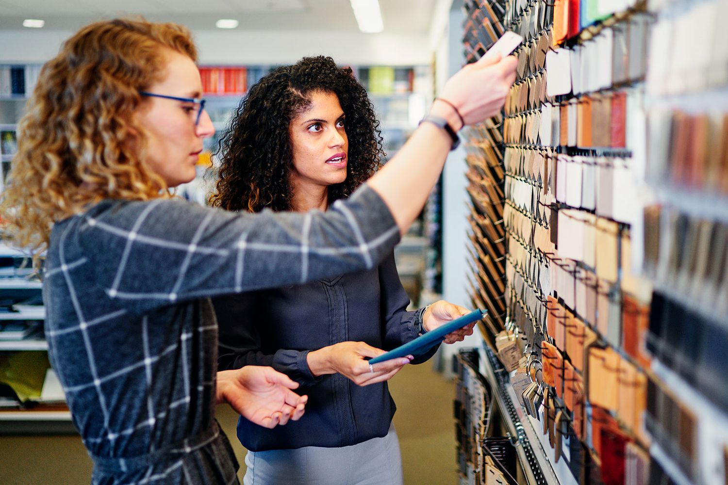 Two women selecting material samples from a wall display in a design or architecture showroom.