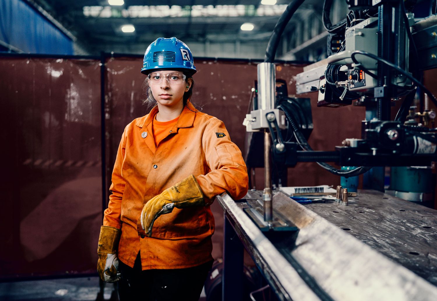 Female worker in orange uniform and blue hard hat operating machinery in manufacturing facility.