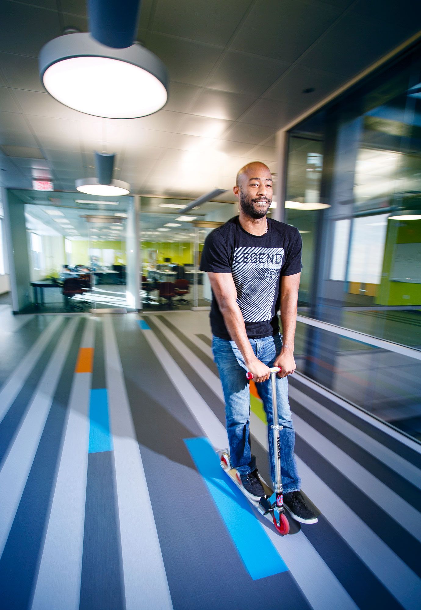 Smiling man rides a scooter through a vibrant modern office hallway with colorful floor stripes.