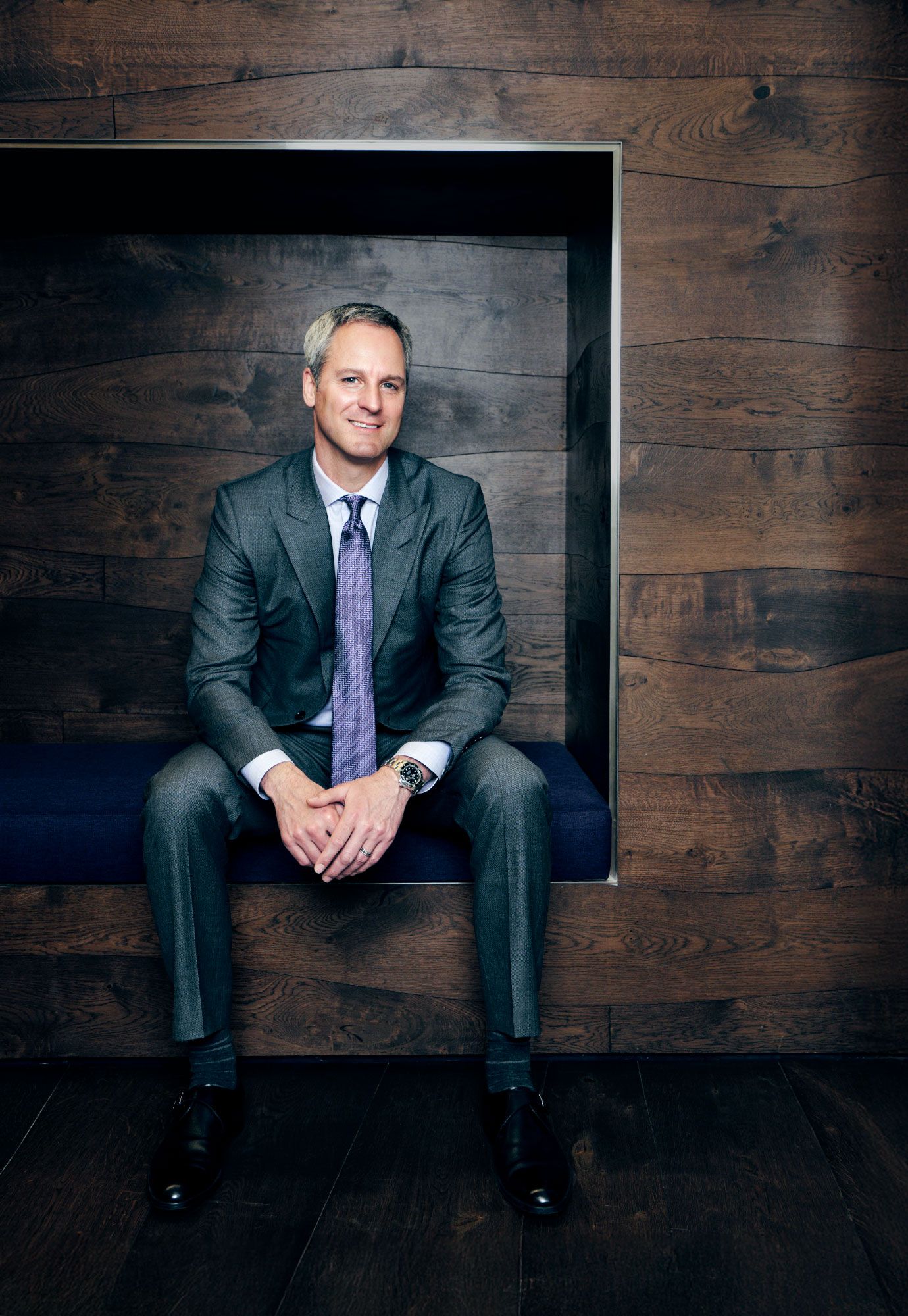 Professional businessman in gray suit sits relaxed on bench against rustic wood panel wall.