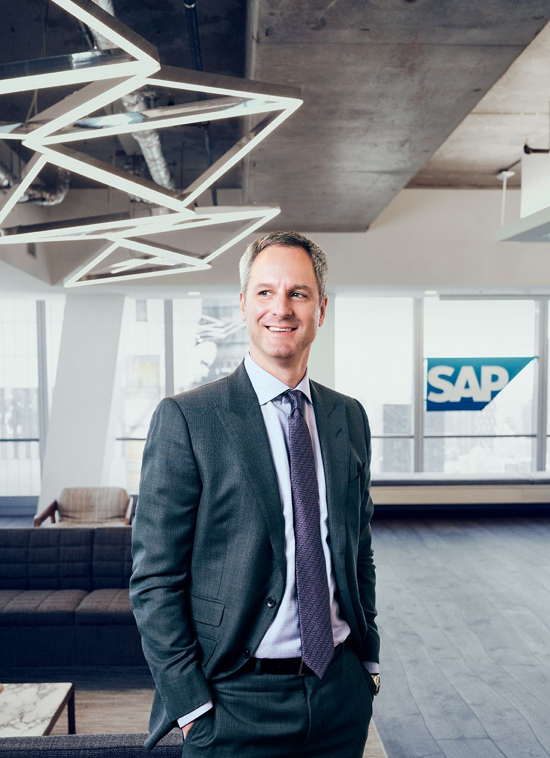 Professional man in gray suit smiling confidently in modern SAP office lobby with geometric lighting fixture.