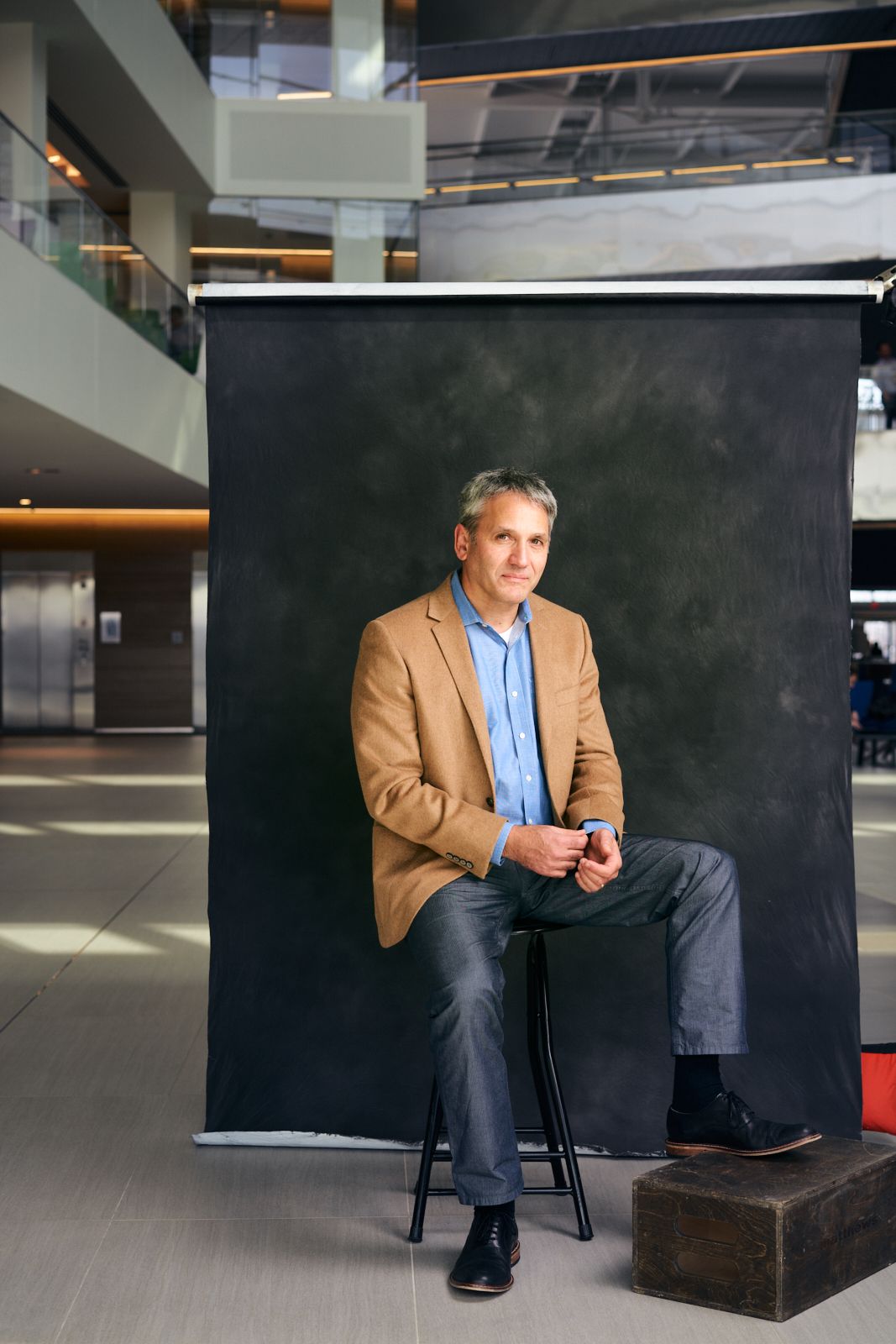 Middle-aged man in tan blazer sits on stool before dark backdrop in modern building interior.