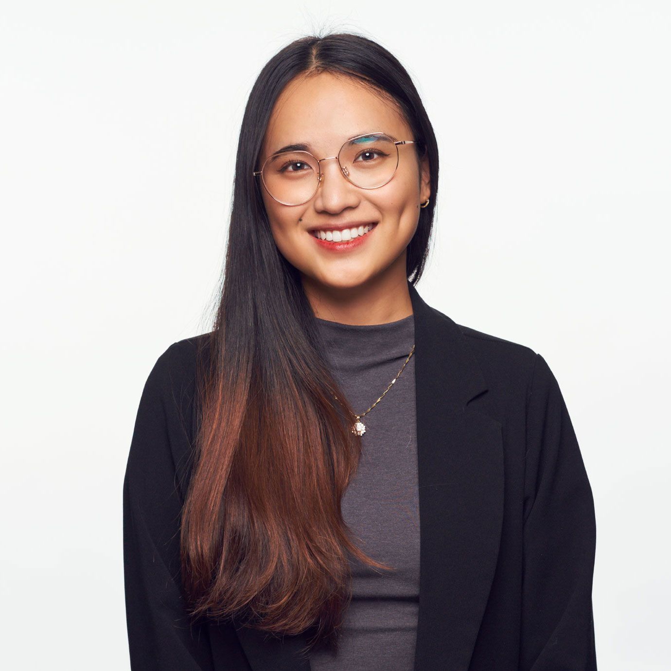 Smiling young woman with long dark hair, glasses, wearing a black blazer and gray top, professional headshot on white background.