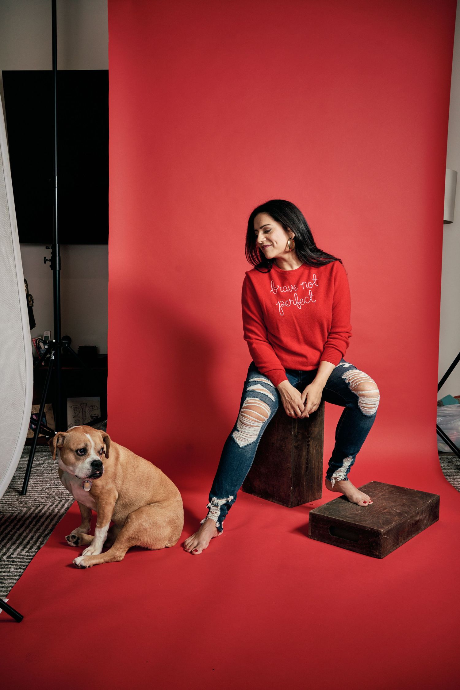 Woman in red sweatshirt and ripped jeans sits on wooden block beside brown dog against red studio backdrop.