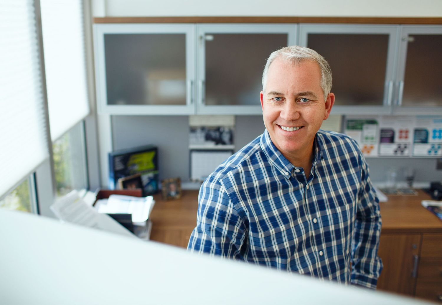 Smiling middle-aged man in plaid shirt sitting at office desk with shelves in background.