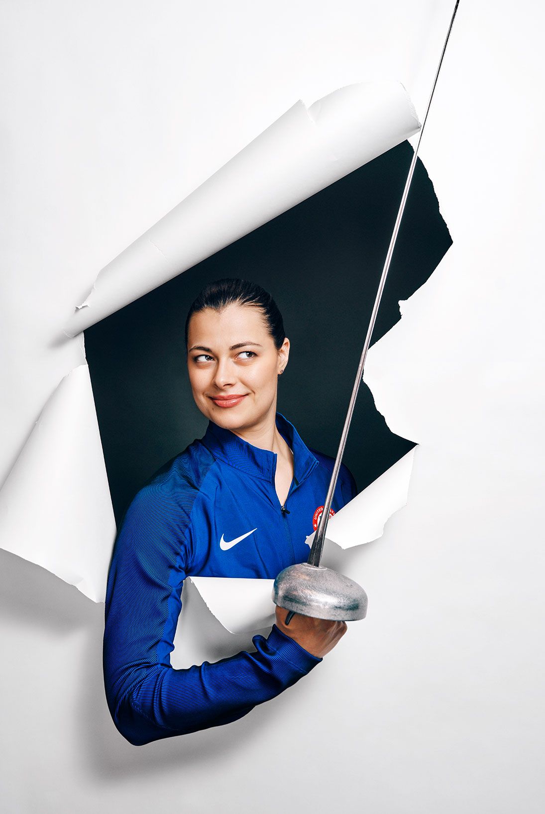 Female fencer in blue Nike jacket holding sword emerging through torn white paper backdrop.