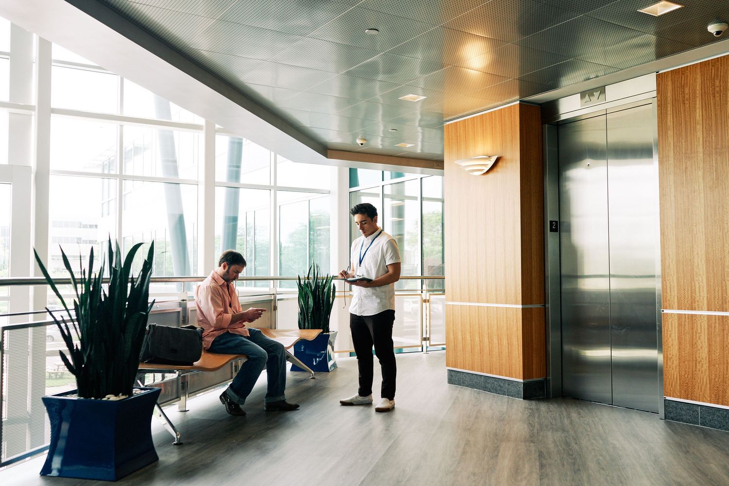 Two professionals converse in a modern office lobby with large windows, plants, and a sleek elevator.