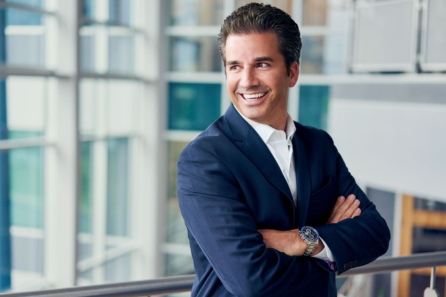 Smiling businessman in navy suit with arms crossed standing in modern office building.