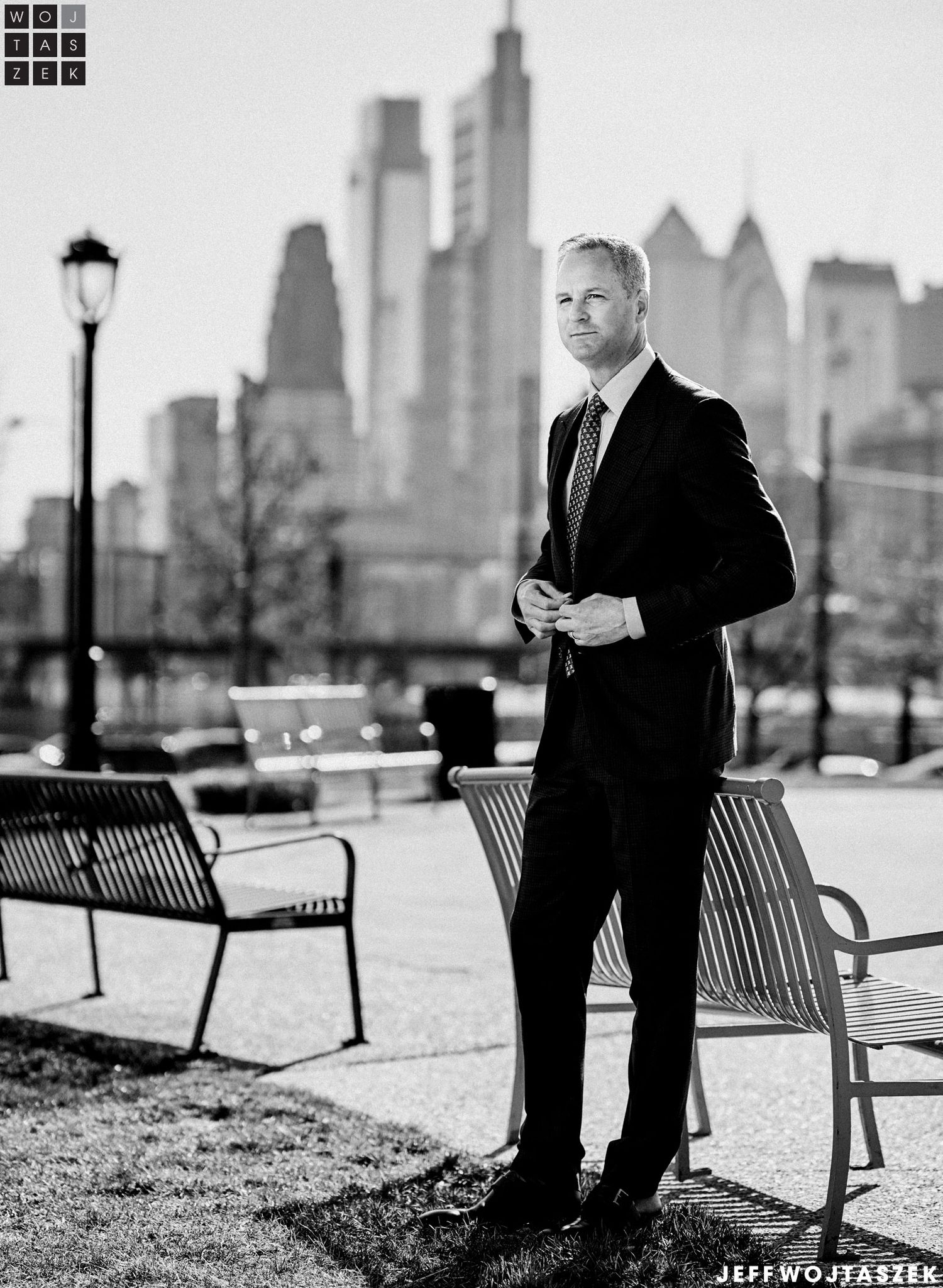 Black and white photo of a man in a suit standing near park benches with a city skyline in the background.