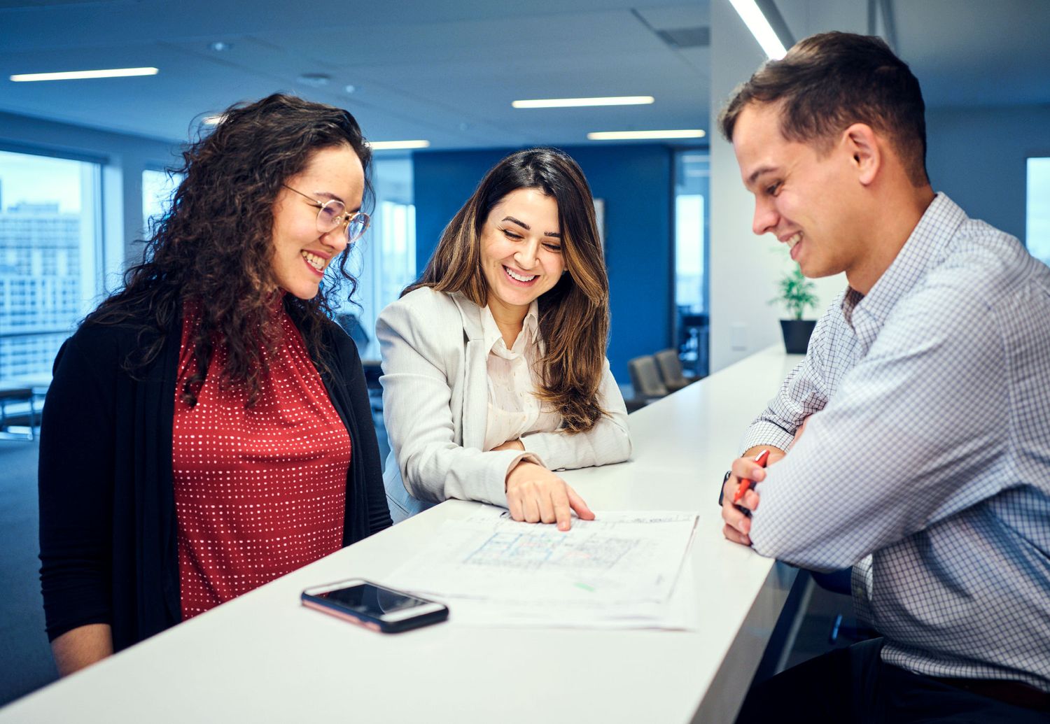 Three diverse colleagues reviewing documents together and smiling at a modern office workspace.