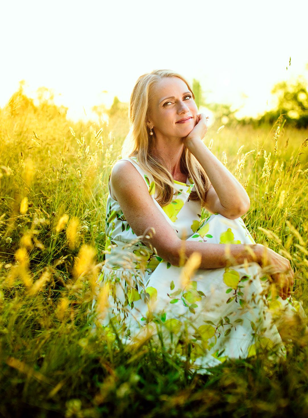 Blonde woman in floral dress sitting peacefully in golden sunlit meadow field.