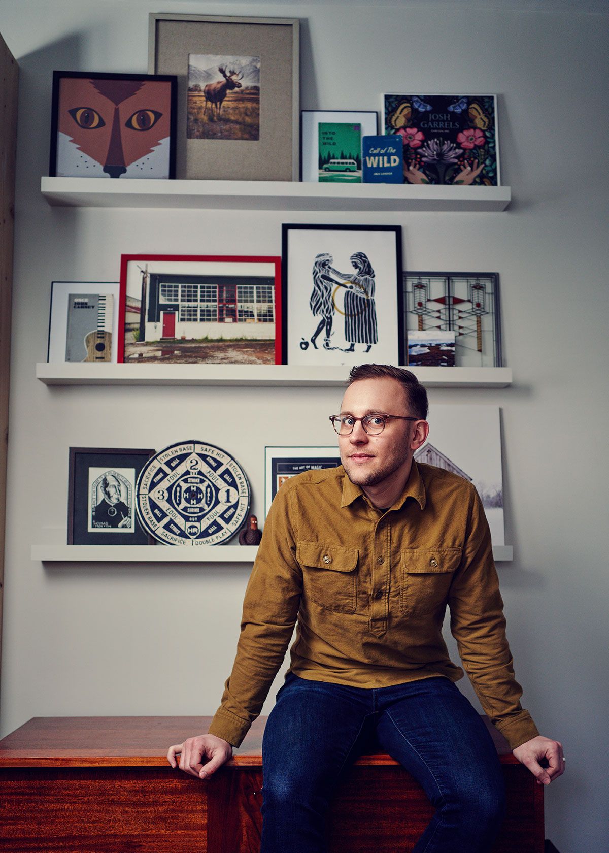 Man with glasses in mustard jacket sits before wall shelves displaying eclectic art prints and framed photographs.