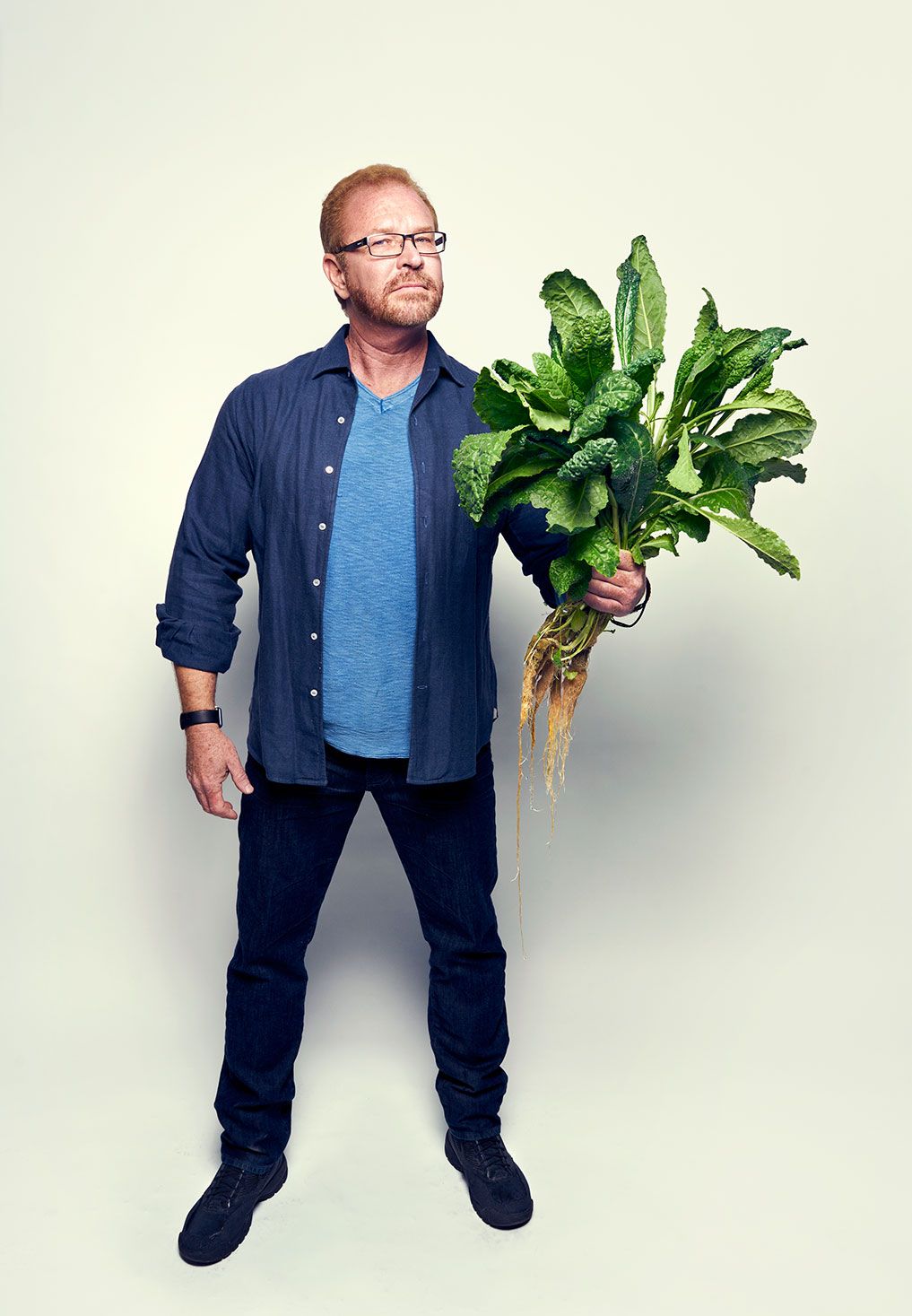 Man in blue shirt holding large fresh leafy green vegetable plant on white background.