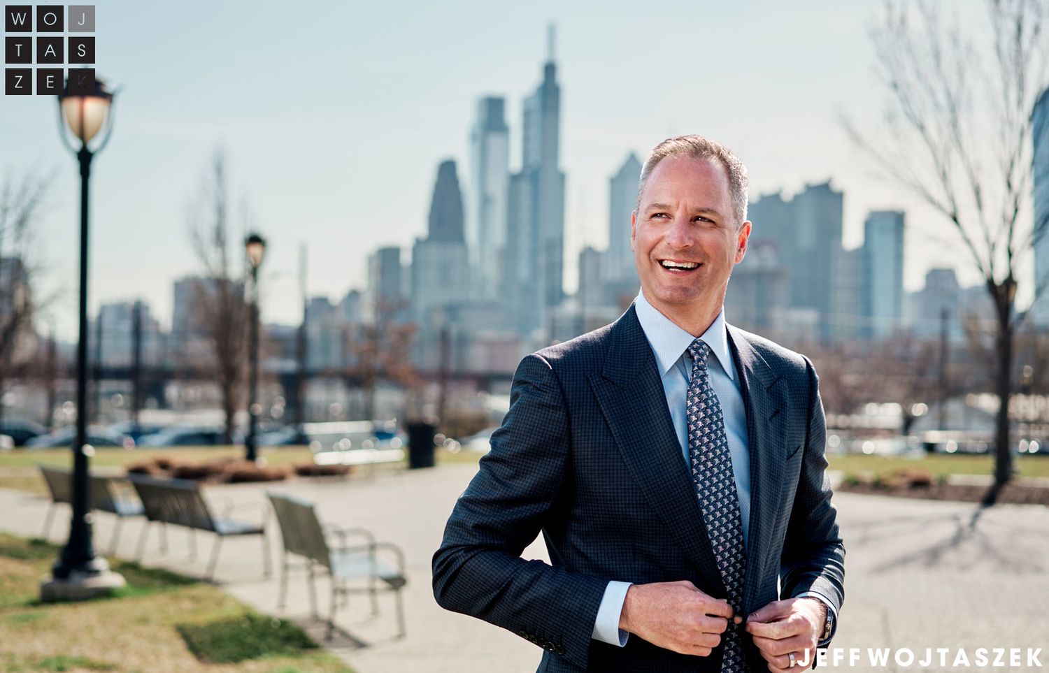 Professional man in navy suit smiling outdoors with Philadelphia city skyline in background on sunny day.