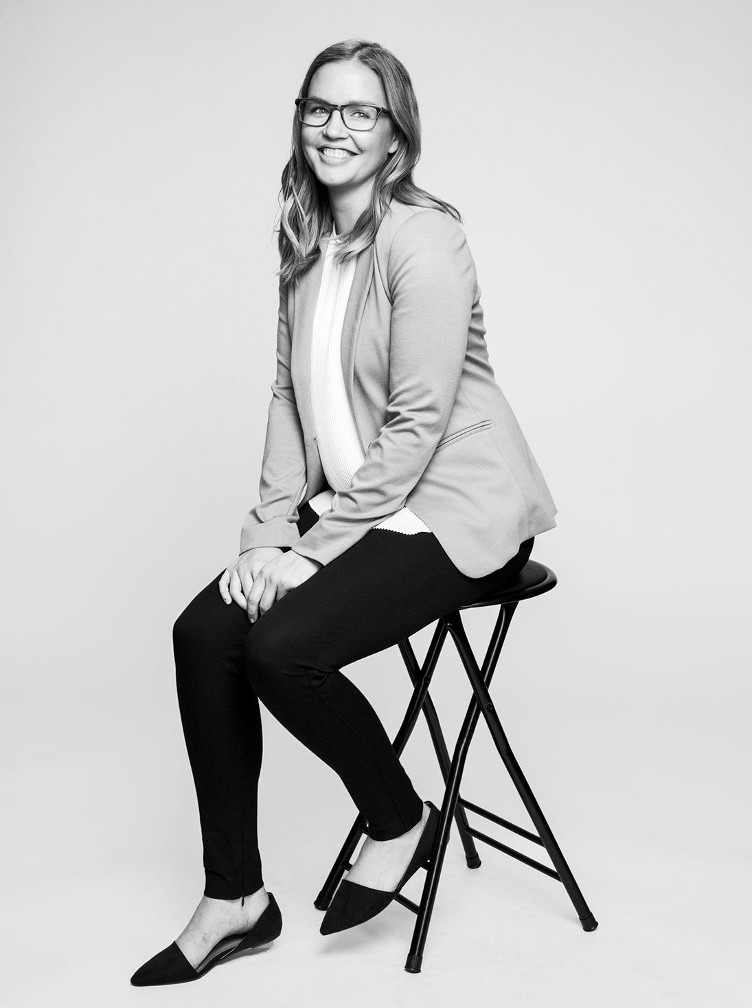 Black and white professional portrait of a smiling woman in blazer and glasses seated on a stool.