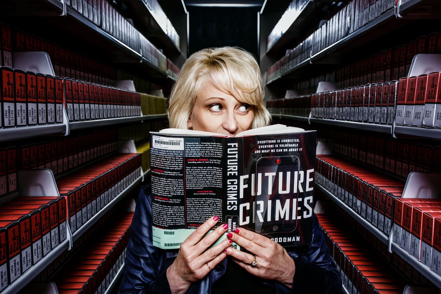 Blonde woman reading Future Crimes book surrounded by rows of red binders in library.