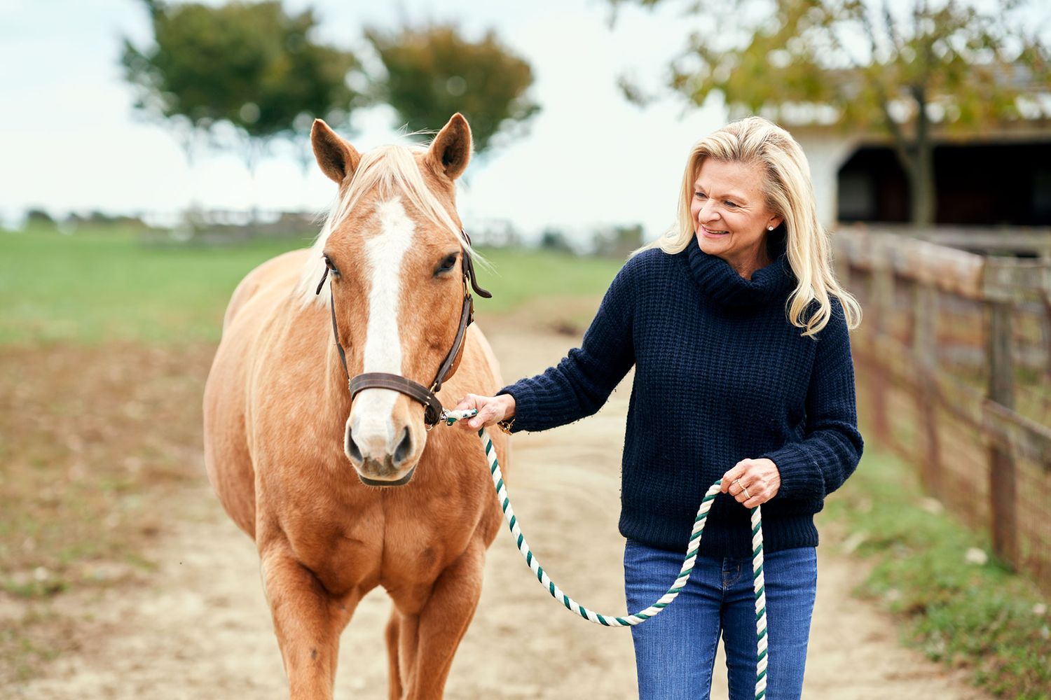 Smiling blonde woman walks a palomino horse along a sunny farm path wearing a navy sweater.
