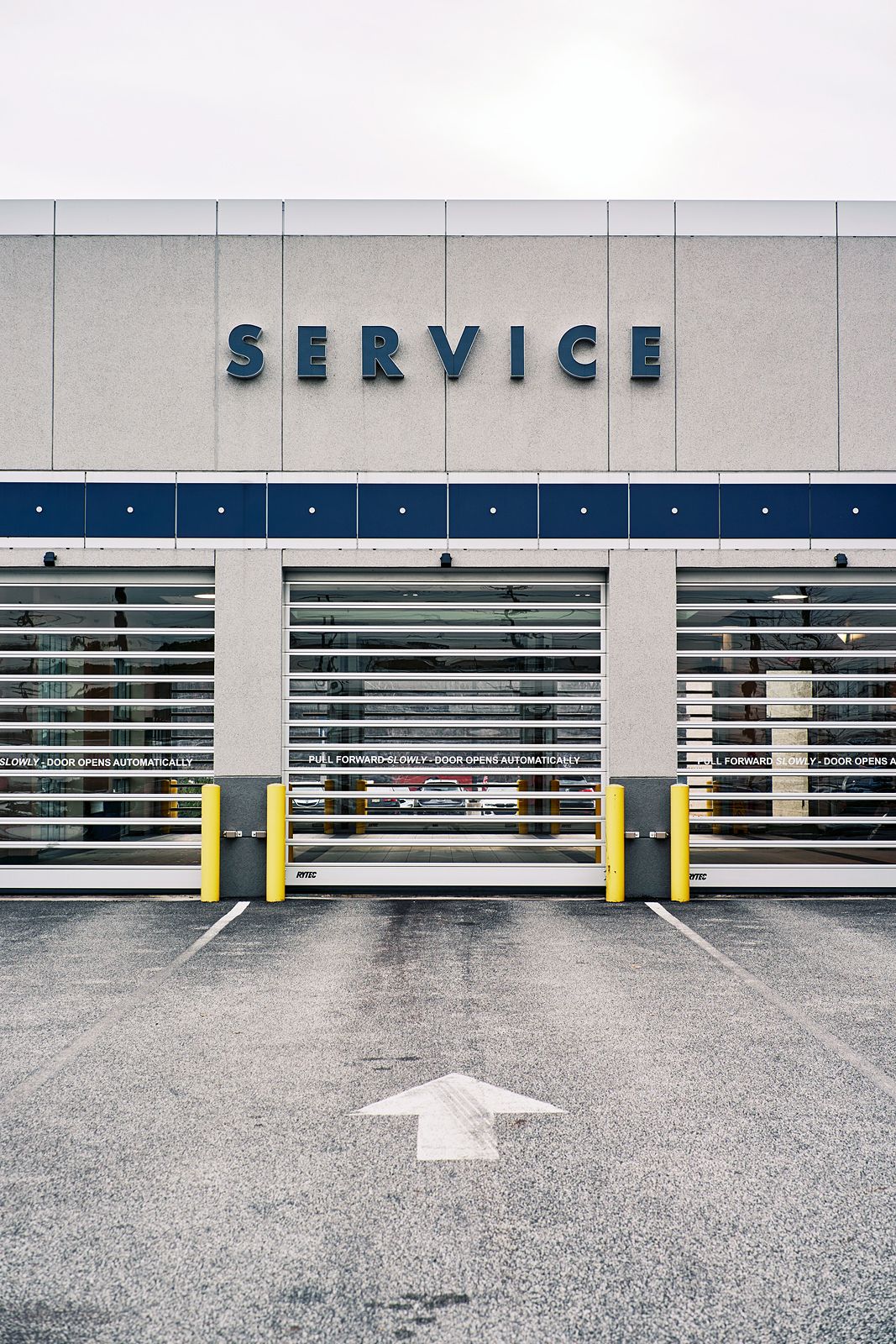 Automotive service center with rolling garage doors, yellow bollards, and a large SERVICE sign on a gray commercial building.