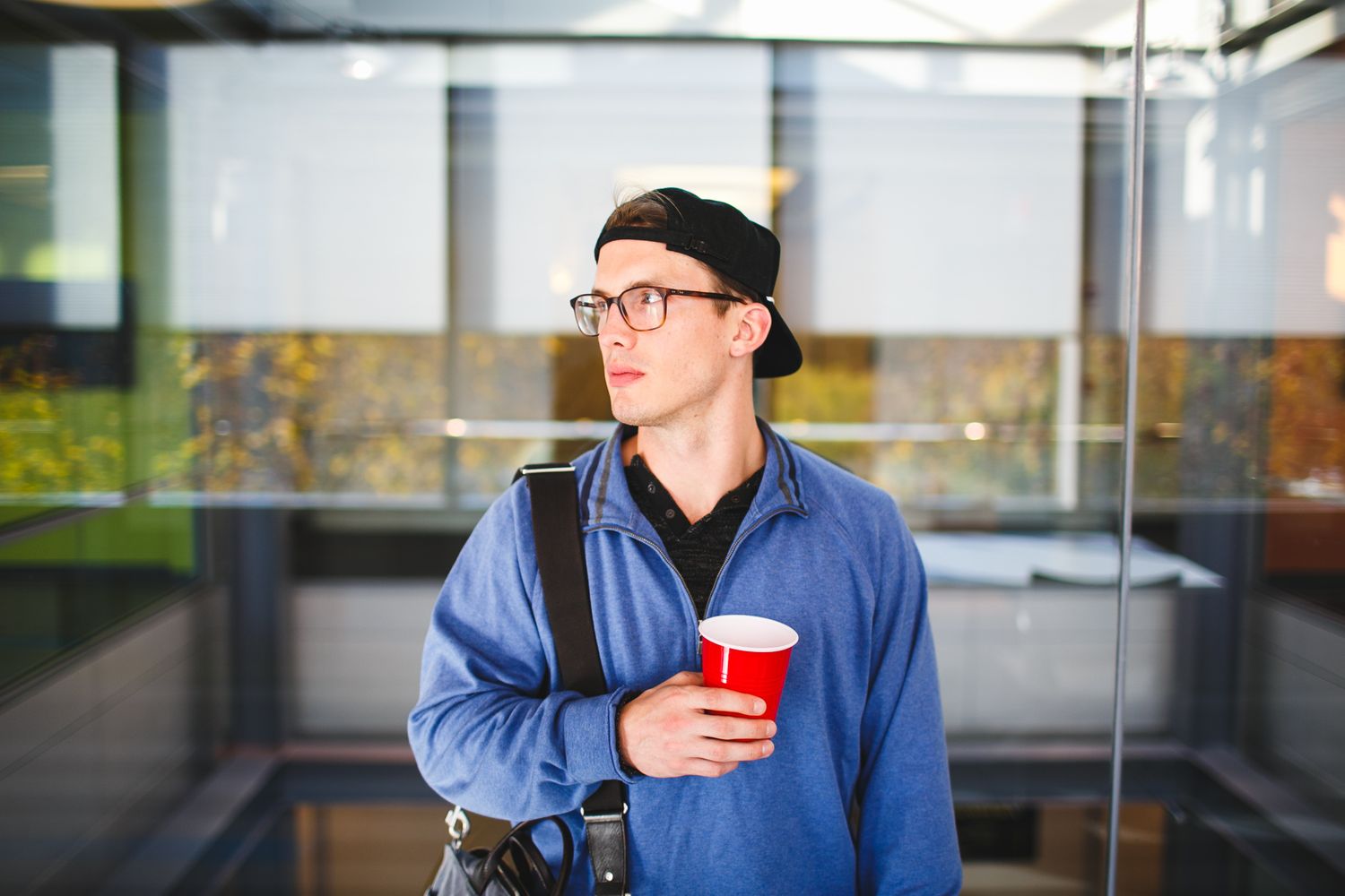 Young man wearing glasses and black cap holding red cup outdoors near glass building.