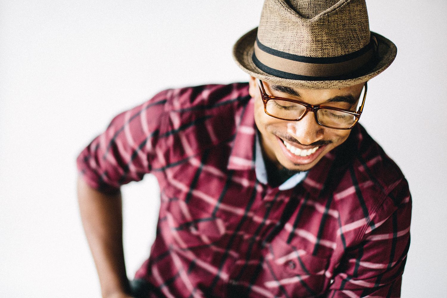 Smiling young man wearing brown fedora hat and glasses in red plaid shirt on white background.