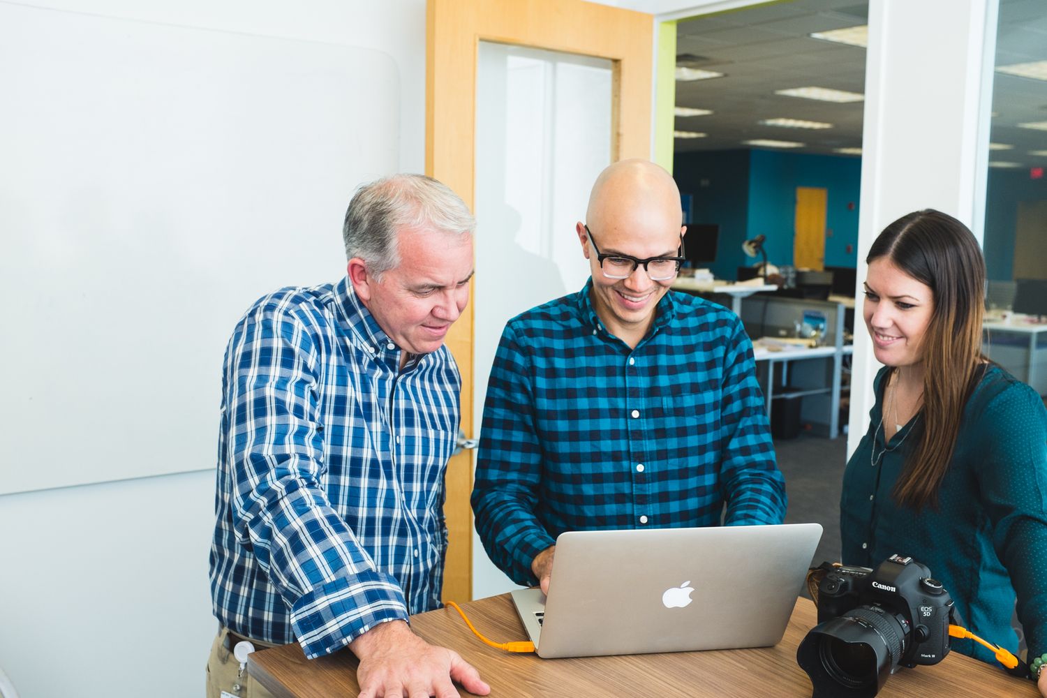 Three colleagues collaborating around a laptop in a modern office, with a camera on the table nearby.