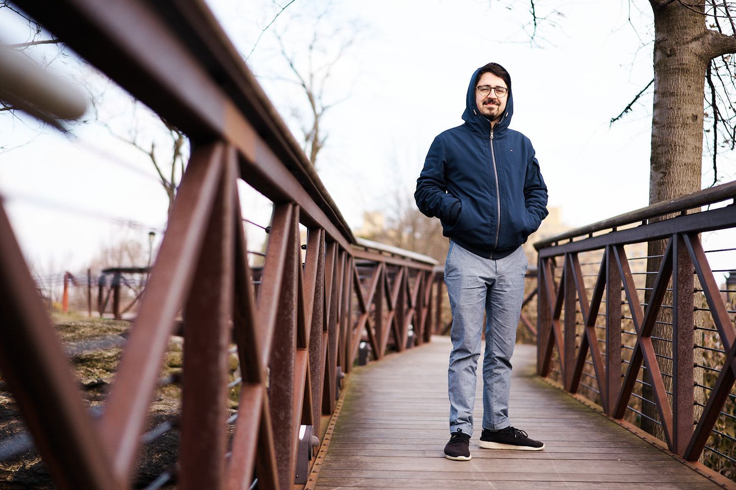 Man in blue hoodie jacket standing on a wooden bridge with red railings surrounded by bare trees.