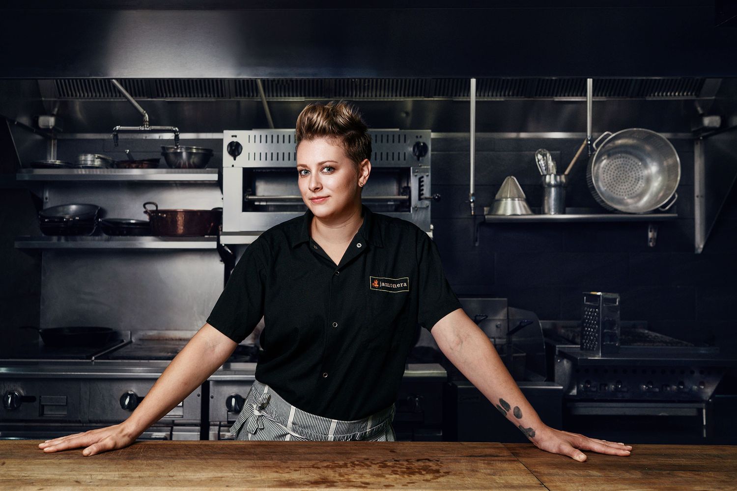 Female chef leaning on wooden prep table in professional commercial kitchen setting.