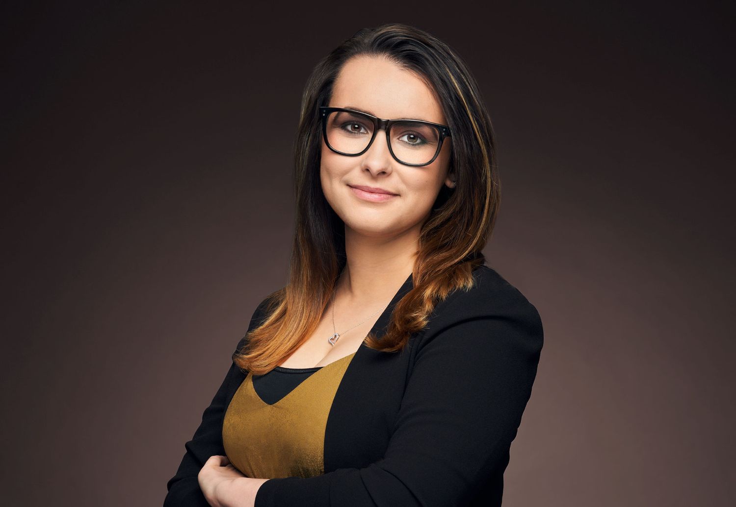 Professional woman with glasses and dark hair wearing a black blazer posing with arms crossed on dark background.