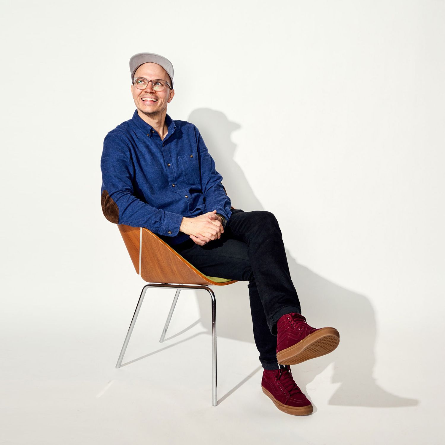 Smiling man in blue shirt and gray beanie sitting relaxed in a modern wooden chair against white background.