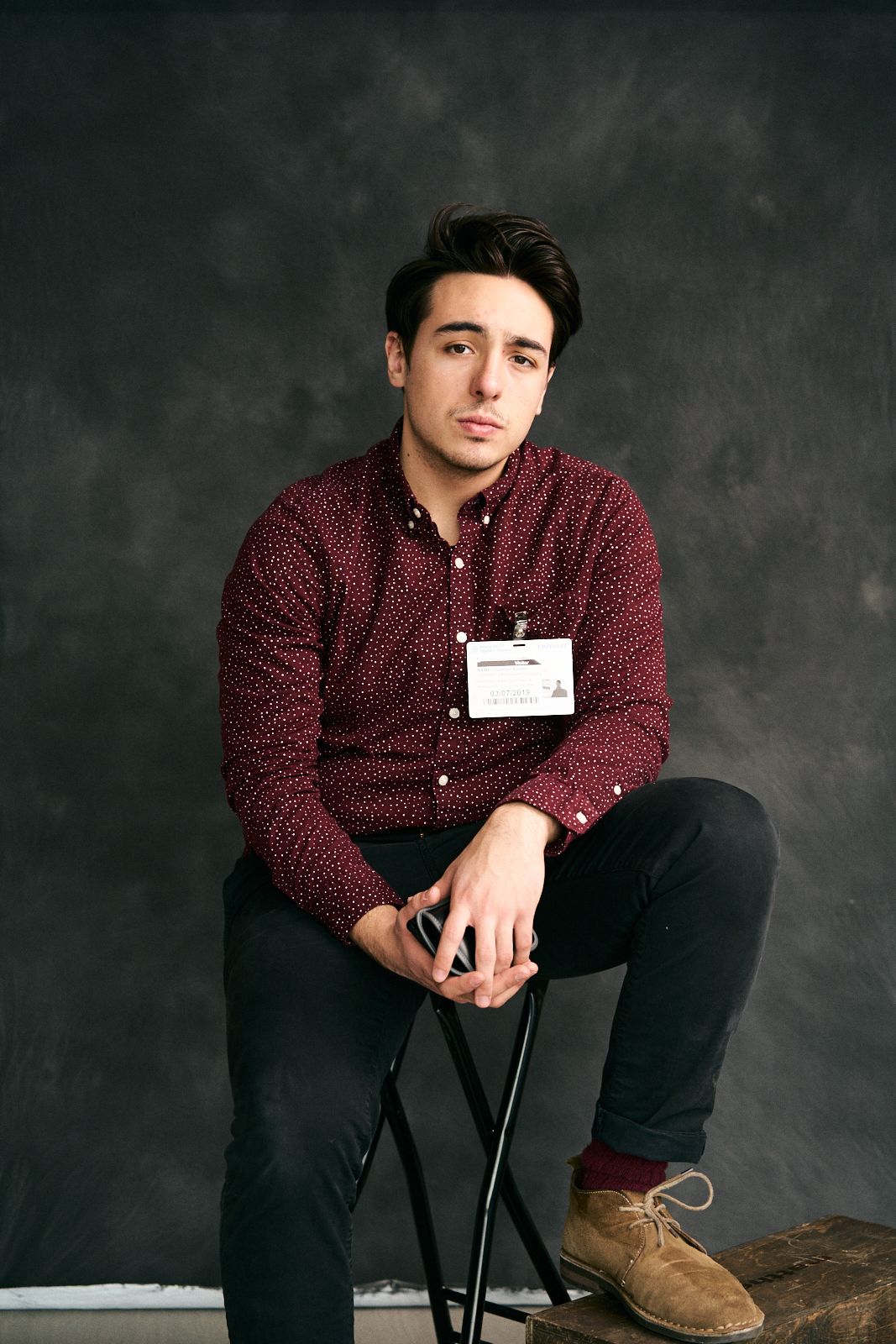 Young man in burgundy patterned shirt sitting on stool against gray backdrop wearing black jeans and tan shoes.