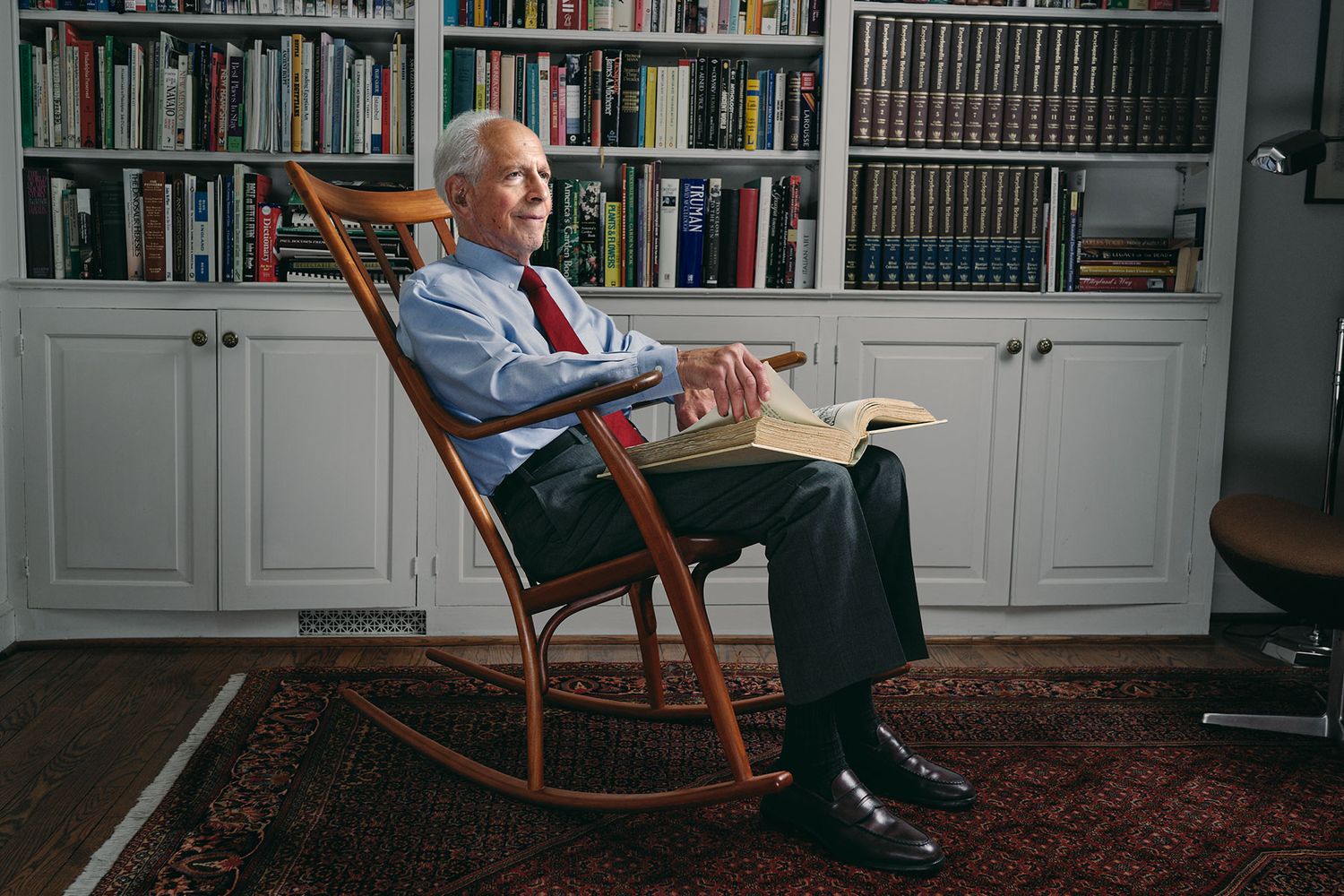 Senior businessman reading book in wooden rocking chair beside built-in library bookshelves.