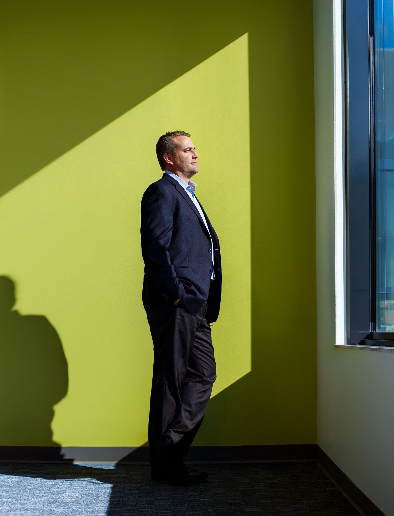 Businessman in dark suit stands contemplatively against a bright yellow-green wall in sunlight.