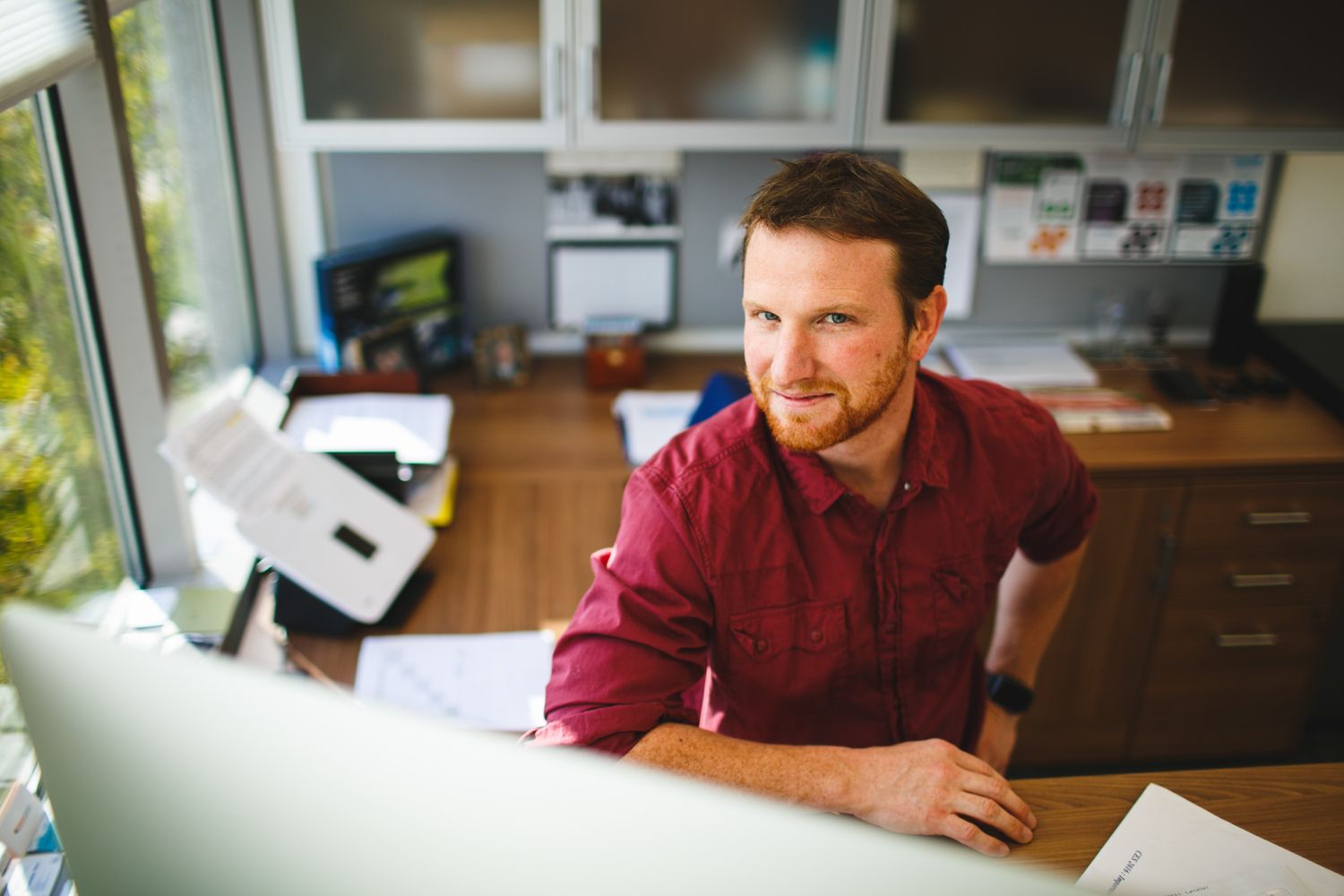 Man in red shirt smiling at desk in modern office workspace with monitors and shelves in background.
