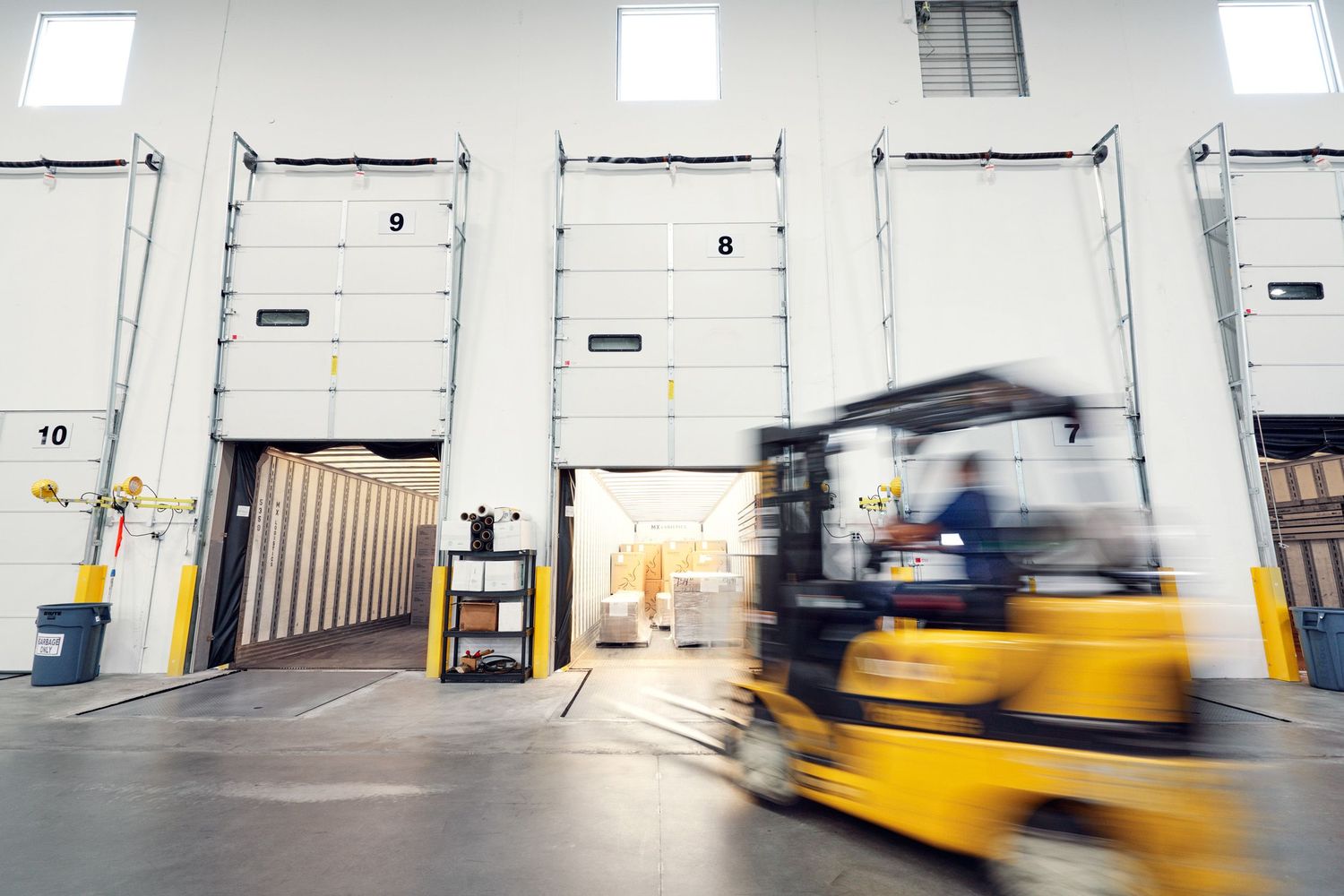 Forklift operator moving equipment in busy industrial warehouse with loading dock doors.