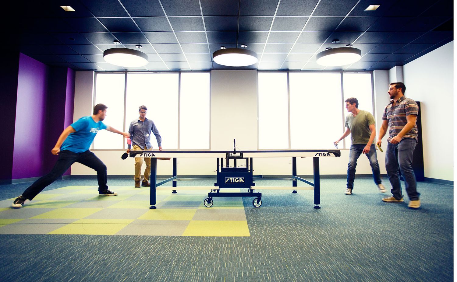 Four colleagues playing ping pong on a Stiga table in a modern colorful office break room.