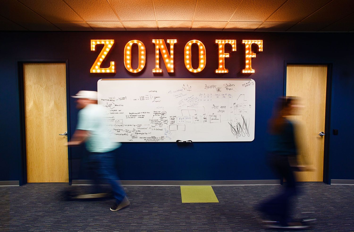 Two people walk past a large whiteboard under illuminated ZONOFF marquee letters in a modern office.