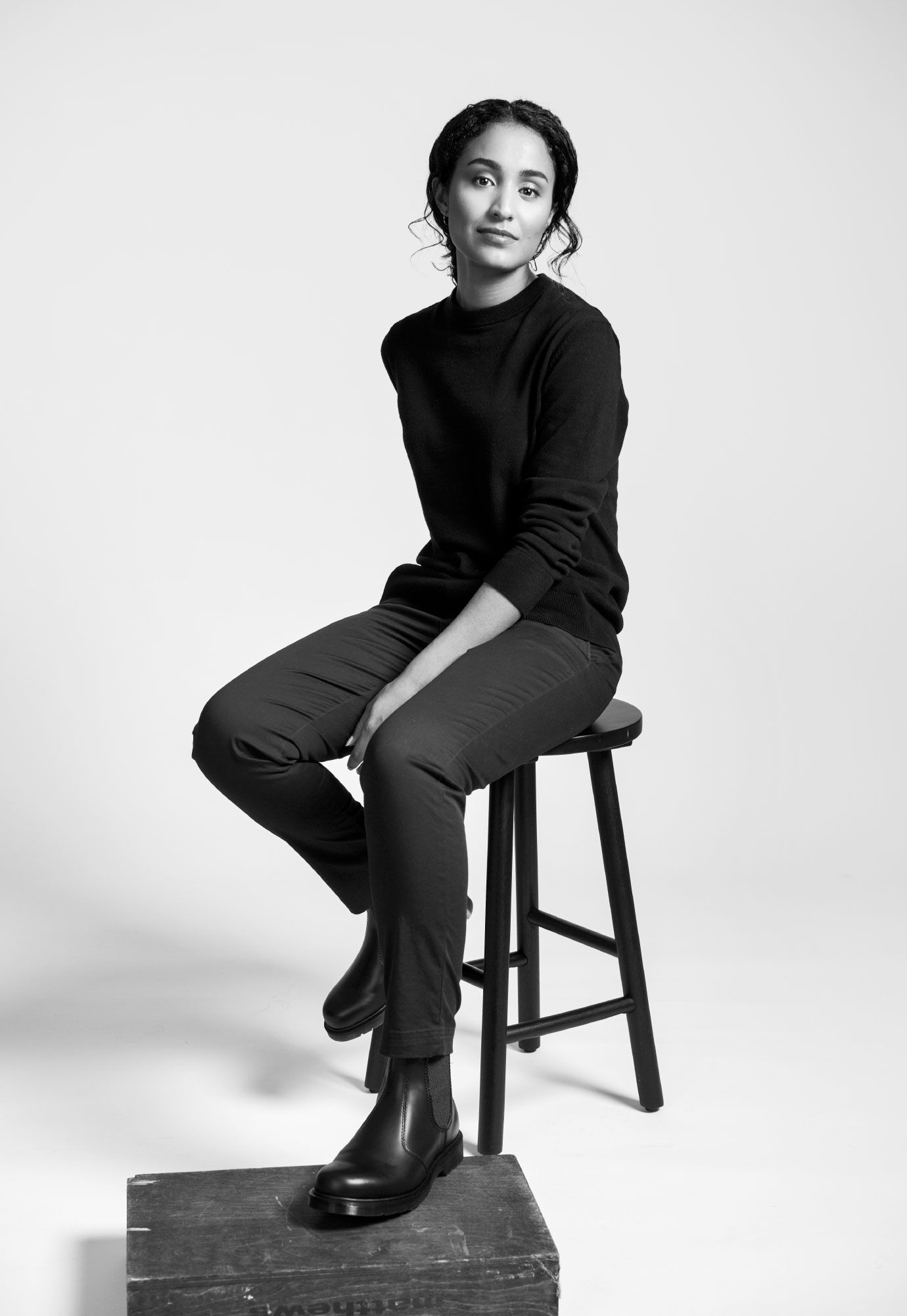 Black and white portrait of a young woman sitting on a stool in casual attire.