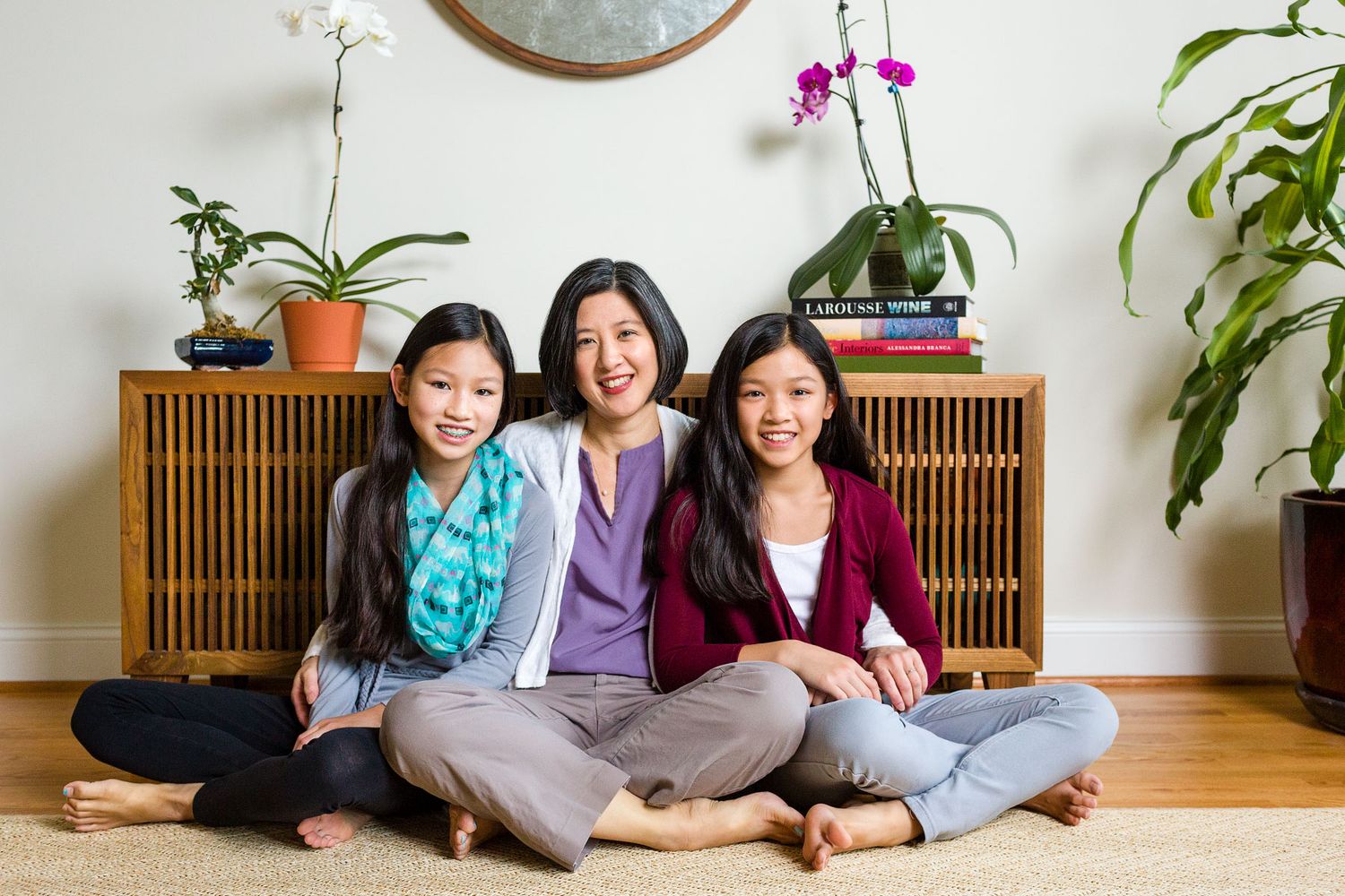 Asian mother and two daughters sitting cross-legged together smiling on living room floor.