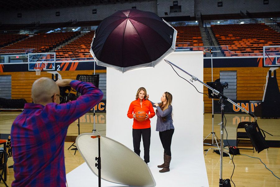 Photographer shoots woman in red hoodie against white backdrop in arena studio setup with umbrella lighting equipment.