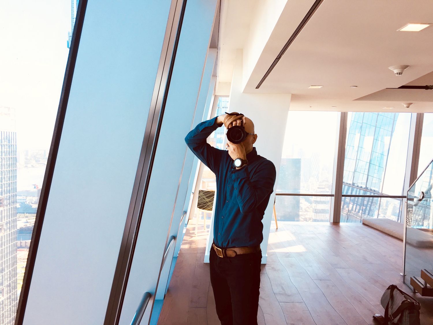 Man in blue shirt gazes at city skyline through floor-to-ceiling windows in a modern high-rise office.
