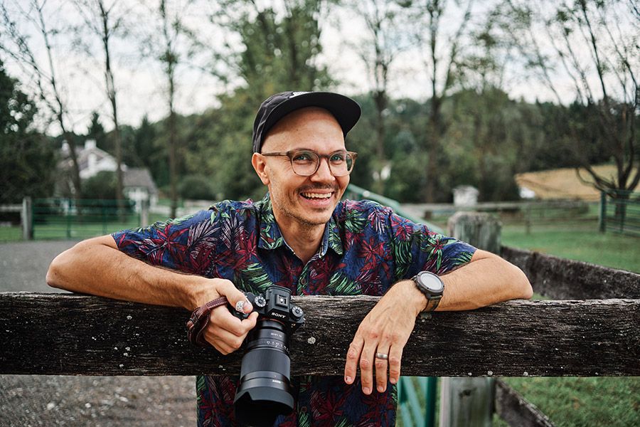 Smiling photographer holding camera while leaning on a wooden fence outdoors.