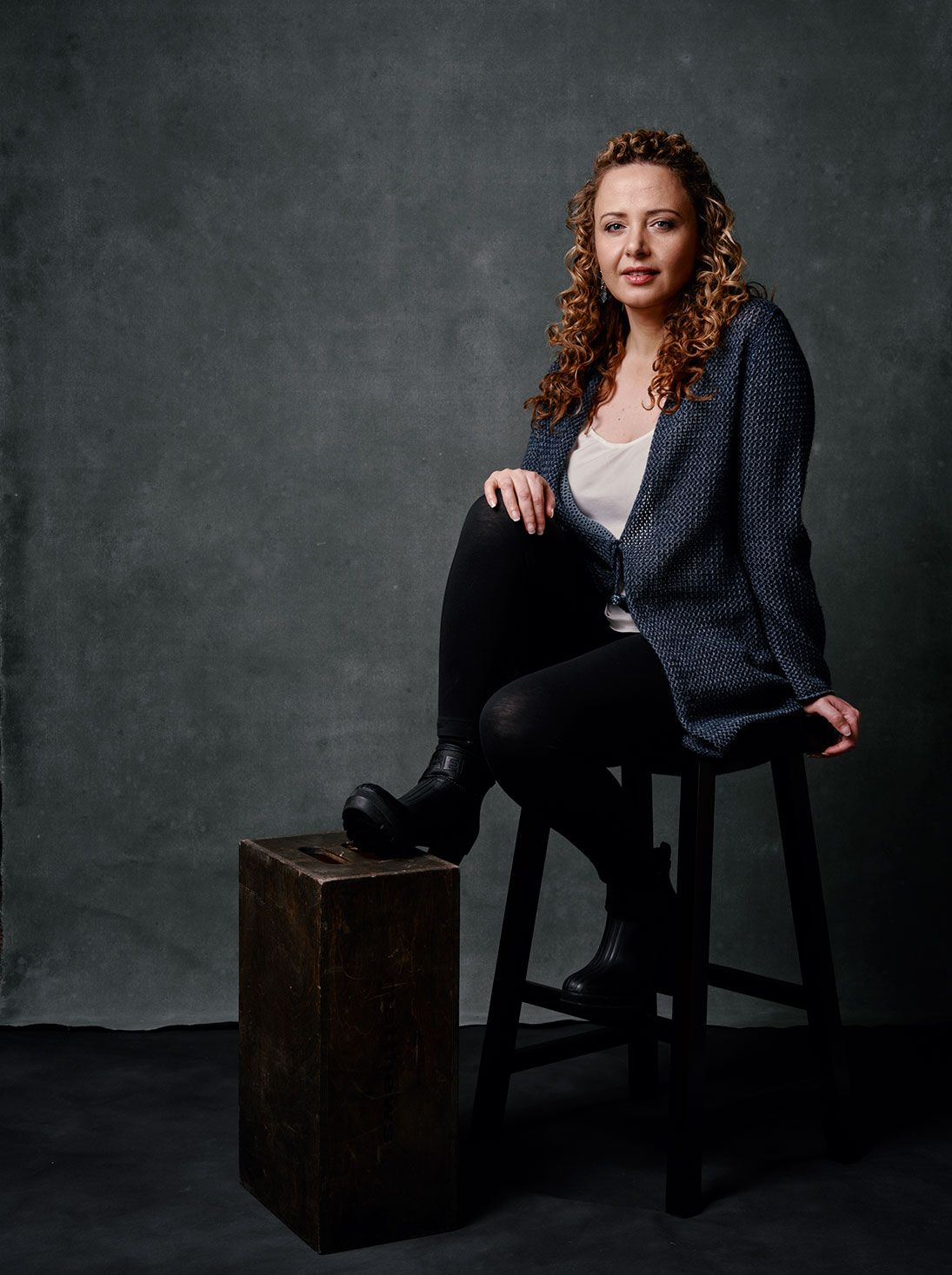 Woman with curly hair sits on stool in blue cardigan and black leggings for professional portrait.