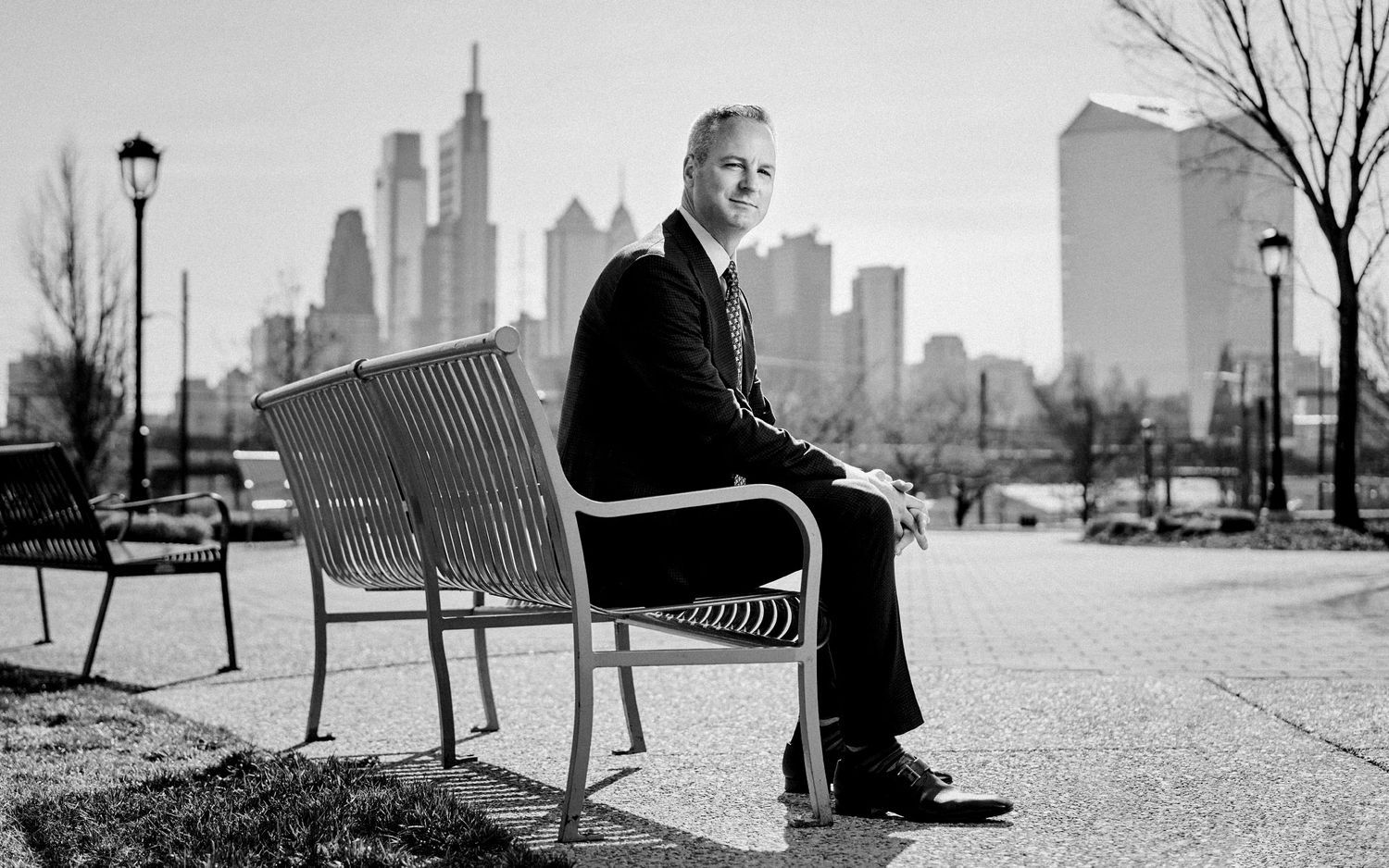 Black and white portrait of businessman seated on bench with Philadelphia city skyline in background.