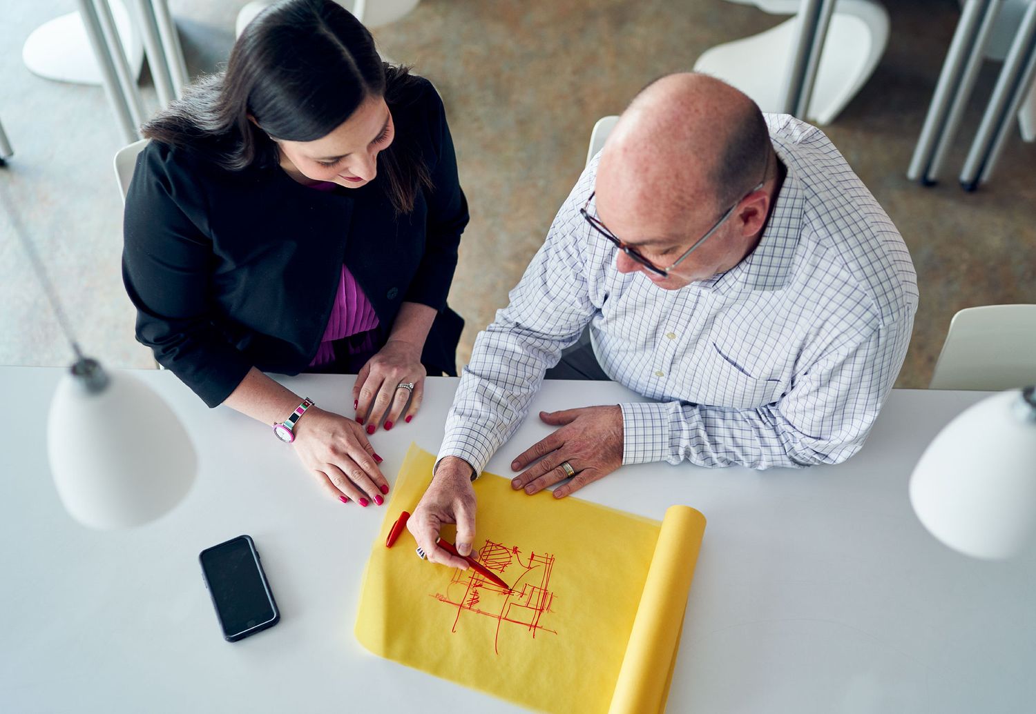 Two professionals review architectural blueprints on yellow paper at a modern white office table.