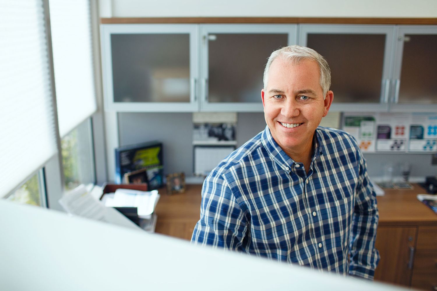 Smiling middle-aged man in plaid shirt sitting at office desk with shelves and computer in background.