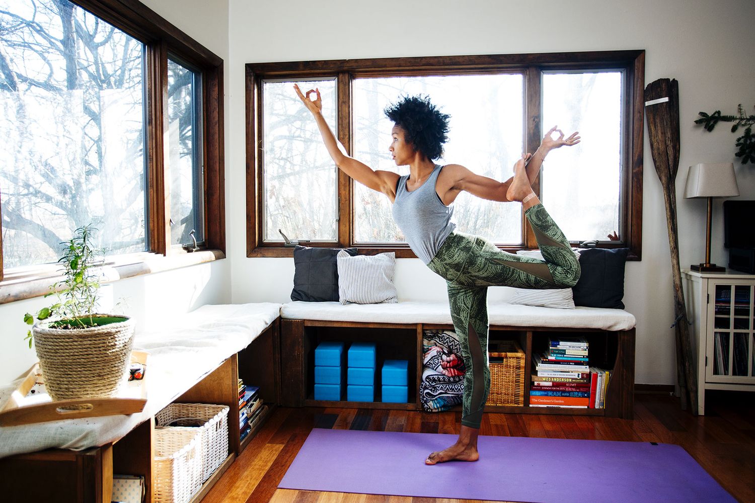 Woman practicing yoga tree pose on purple mat in bright home studio with large windows.