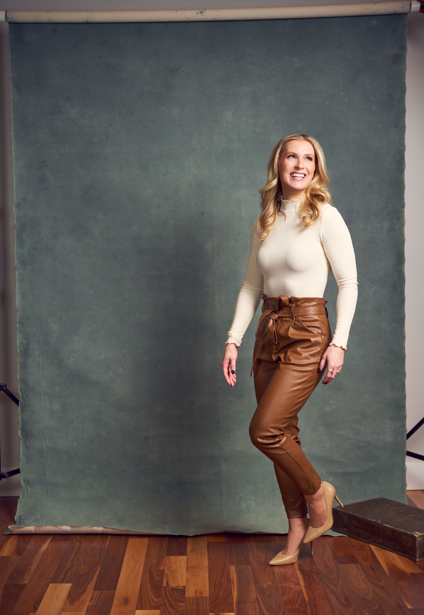 Smiling blonde woman in cream turtleneck and brown leather pants poses against gray backdrop.